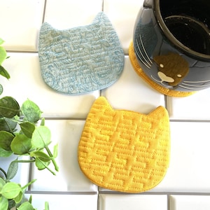 May include: Three cat-shaped fabric coasters. One is blue with a paisley pattern, one is yellow with a geometric pattern, and one is black with white whiskers.