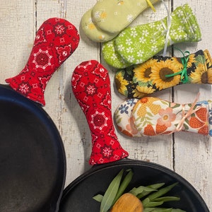 May include: A collection of colorful cast iron skillet handle covers in various floral and patterned fabrics. The covers are red, green, yellow, and floral, with a cast iron skillet in the foreground.