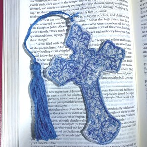 May include: Blue embroidered cross bookmark with a tassel, resting on an open book. The cross features a floral design and white trim. The bookmark is a religious item.