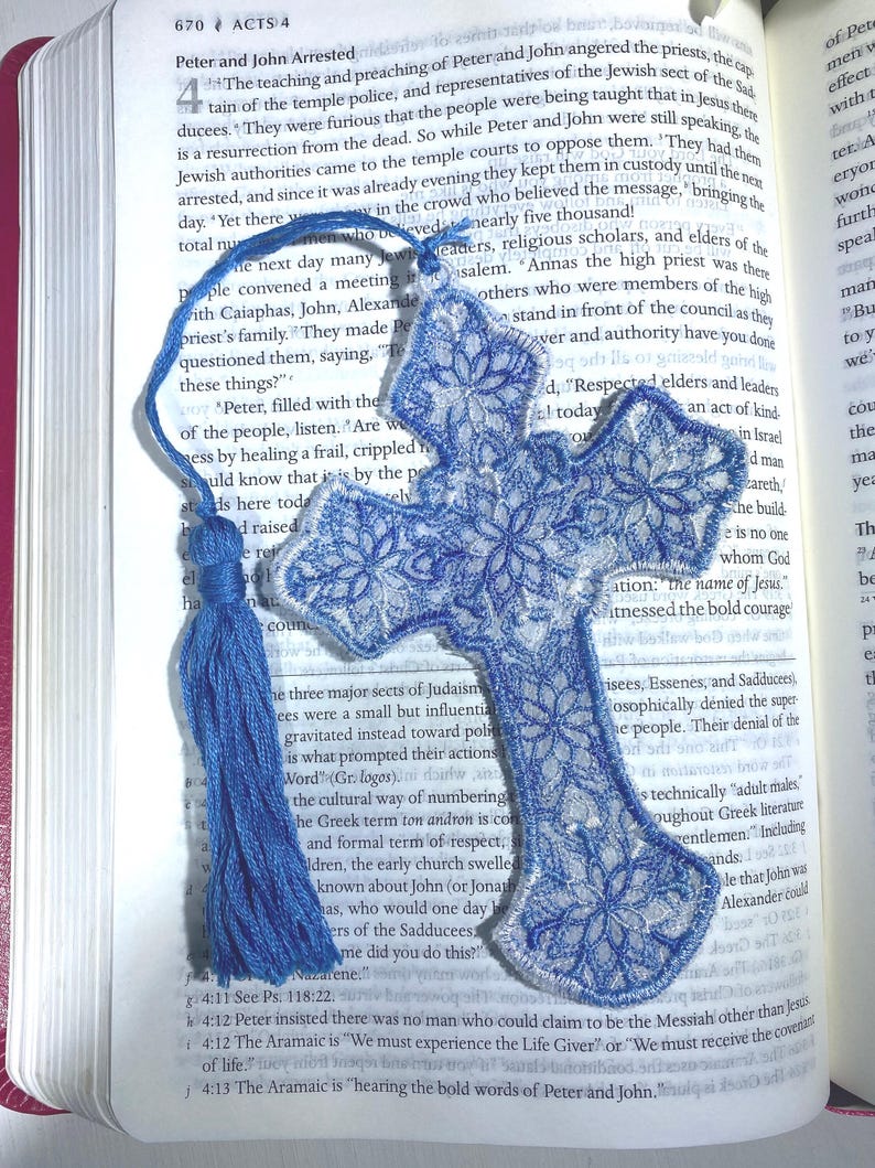 May include: Blue embroidered cross bookmark with a tassel, resting on an open book. The cross features a floral design and white trim. The bookmark is a religious item.