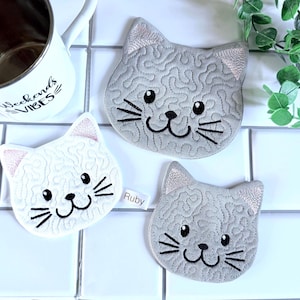 May include: Three cat-shaped coasters in shades of gray and white, with embroidered details. The coasters feature black whiskers, eyes, and smiles. One coaster has a pink ear. A white mug with the text "Weekends Vibes" is visible.