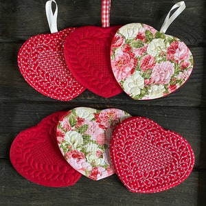 May include: Six red heart-shaped quilted hot pads with different designs. Some have floral patterns, others have a quilted pattern with white dots. All have a white loop for hanging.