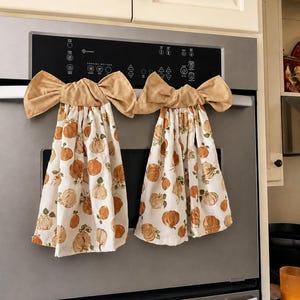 May include: Two hanging kitchen towels featuring a white background with a repeating pattern of orange and tan pumpkins and green leaves. Each towel is topped with a tan, textured fabric bow that secures the towel to a handle.
