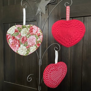 May include: Three heart-shaped quilted ornaments hanging from a decorative metal stand. One heart features a floral pattern, another is solid red with a quilted design, and the third is red with a white dotted pattern.