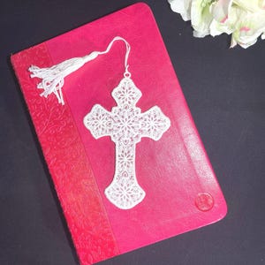 May include: A white, lace cross bookmark with a tassel rests on a closed, red book. The bookmark features an intricate floral design. The book has a textured cover with a floral pattern and a red spine.