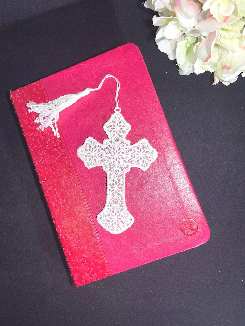 May include: A white, lace cross bookmark with a tassel rests on a closed, red book. The bookmark features an intricate floral design. The book has a textured cover with a floral pattern and a red spine.