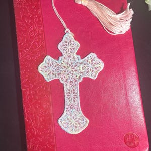 May include: A white, intricately designed cross-shaped bookmark with a delicate floral pattern, resting on a red book. The bookmark has a tassel at the end. The book has the word "BIBLE" on it.