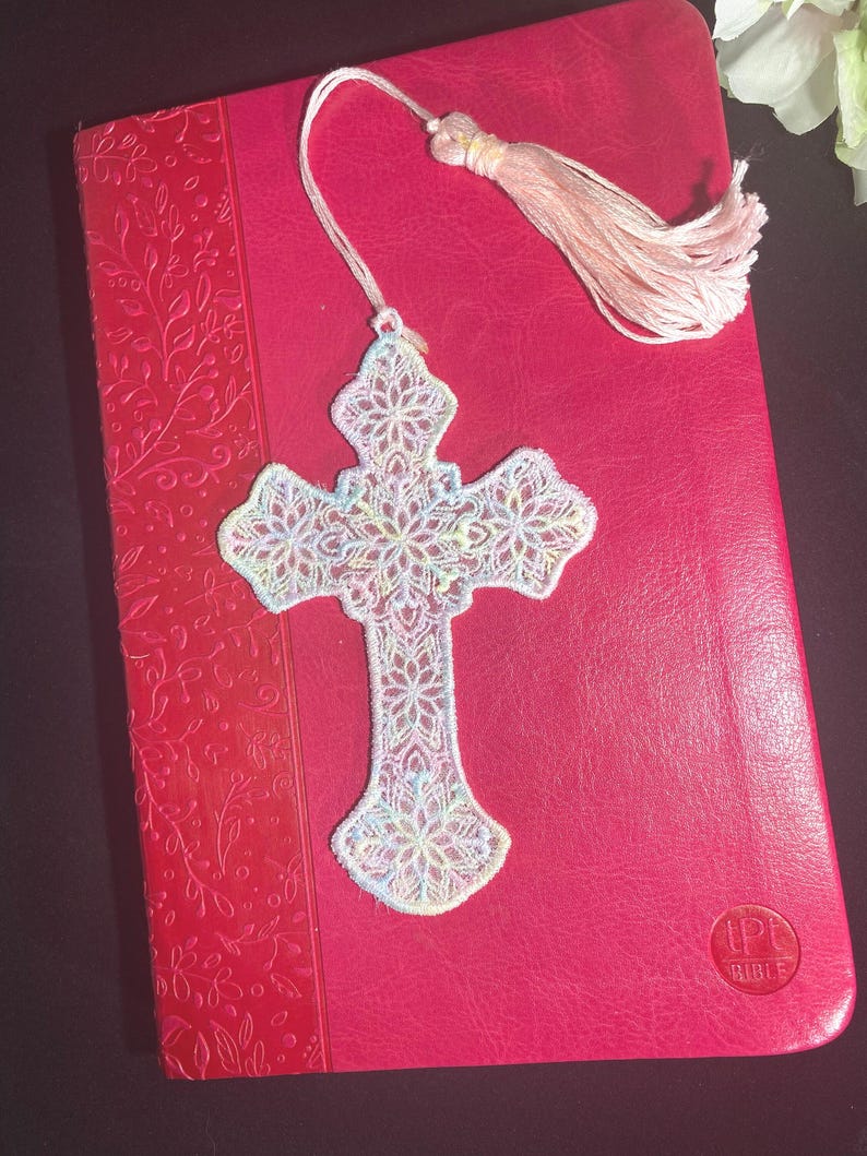 May include: A white, intricately designed cross-shaped bookmark with a delicate floral pattern, resting on a red book. The bookmark has a tassel at the end. The book has the word "BIBLE" on it.