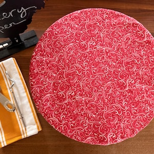 May include: A red and white patterned round fabric cover, possibly for a cake or pie, sits on a wooden surface. An orange and white striped towel with a measuring spoon and whisk are next to it. A chalkboard sign that says "Bakery Open" is also visible.