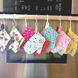 May include: A collection of colorful, patterned oven mitts and pot holders hanging on hooks. The mitts and pot holders feature various designs, including ladybugs, strawberries, and floral patterns. The color palette includes pink, blue, green, and yellow.