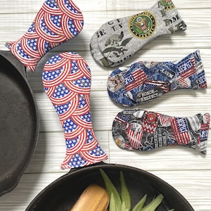 May include: A collection of patriotic-themed cast iron skillet handle covers. Two covers feature a red, white, and blue American flag design. Other covers display military emblems and the words "Honor" and "Respect".
