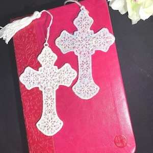 May include: Two white lace cross bookmarks with intricate floral patterns rest on a red book. One bookmark has a white tassel. The crosses are detailed with delicate designs, suggesting a religious or spiritual theme.
