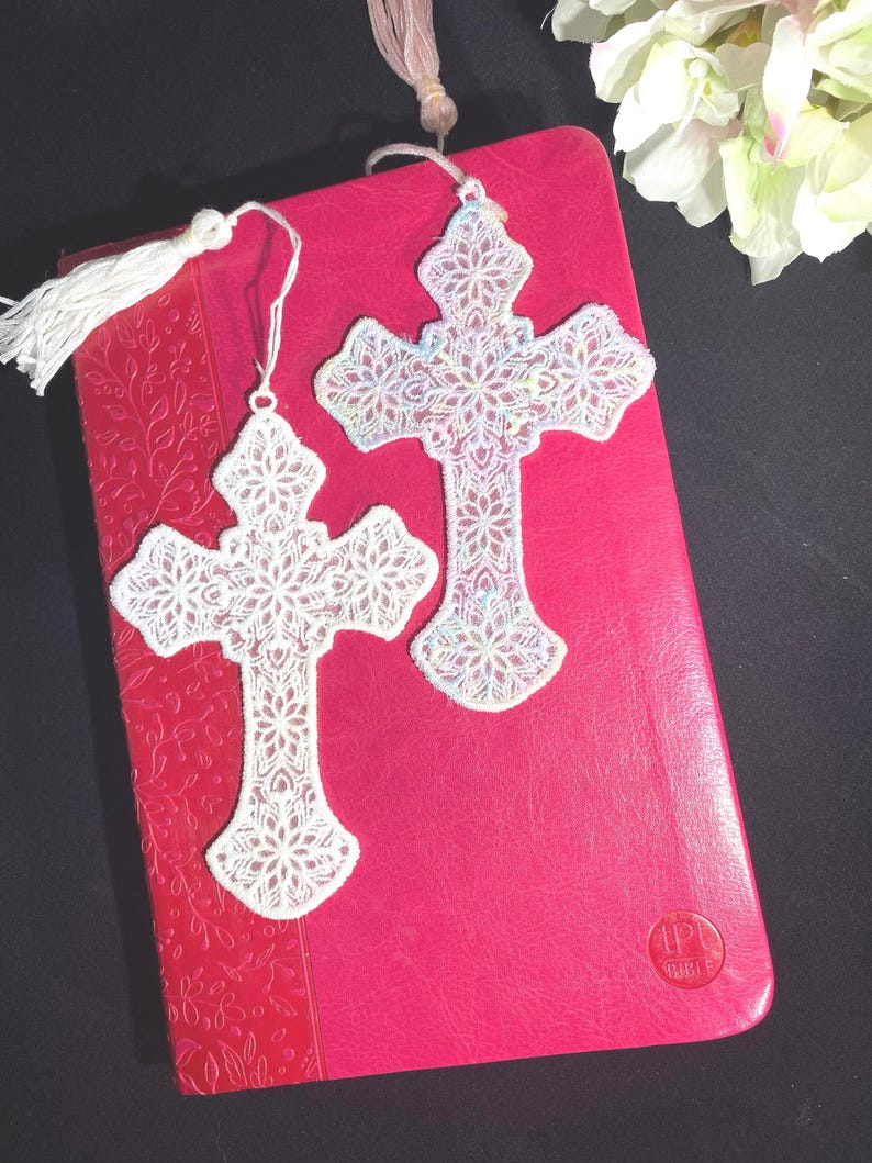 May include: Two white lace cross bookmarks with intricate floral patterns rest on a red book. One bookmark has a white tassel. The crosses are detailed with delicate designs, suggesting a religious or spiritual theme.