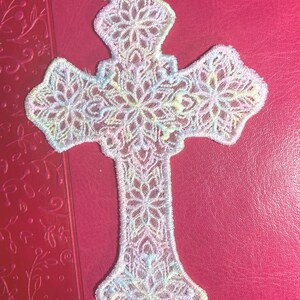 May include: A delicate, openwork cross in pastel colors, featuring intricate floral patterns. The cross is made of a soft, possibly fabric-like material, with a loop at the top for hanging. The color palette includes shades of white, pink, blue, and yellow.