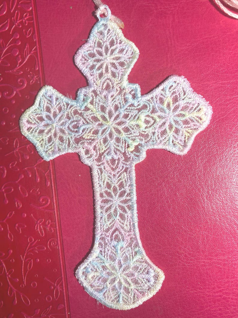 May include: A delicate, openwork cross in pastel colors, featuring intricate floral patterns. The cross is made of a soft, possibly fabric-like material, with a loop at the top for hanging. The color palette includes shades of white, pink, blue, and yellow.