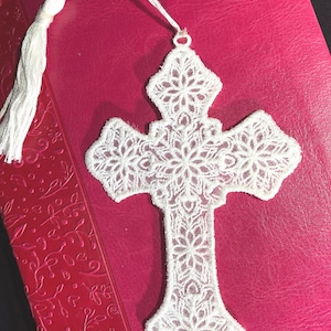 May include: White lace cross bookmark with a tassel, resting on a pink book. The cross features a detailed floral design. The bookmark is made of delicate, textured lace.