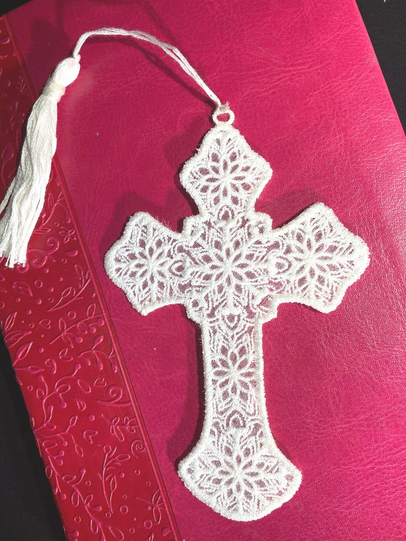 May include: White lace cross bookmark with a tassel, resting on a pink book. The cross features a detailed floral design. The bookmark is made of delicate, textured lace.