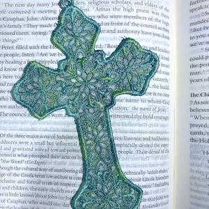 May include: Embroidered teal and green cross-shaped bookmark with floral detailing. The bookmark is made of a delicate, open-weave fabric and has a teal cord for easy placement in a book. The bookmark is resting on the pages of a book.