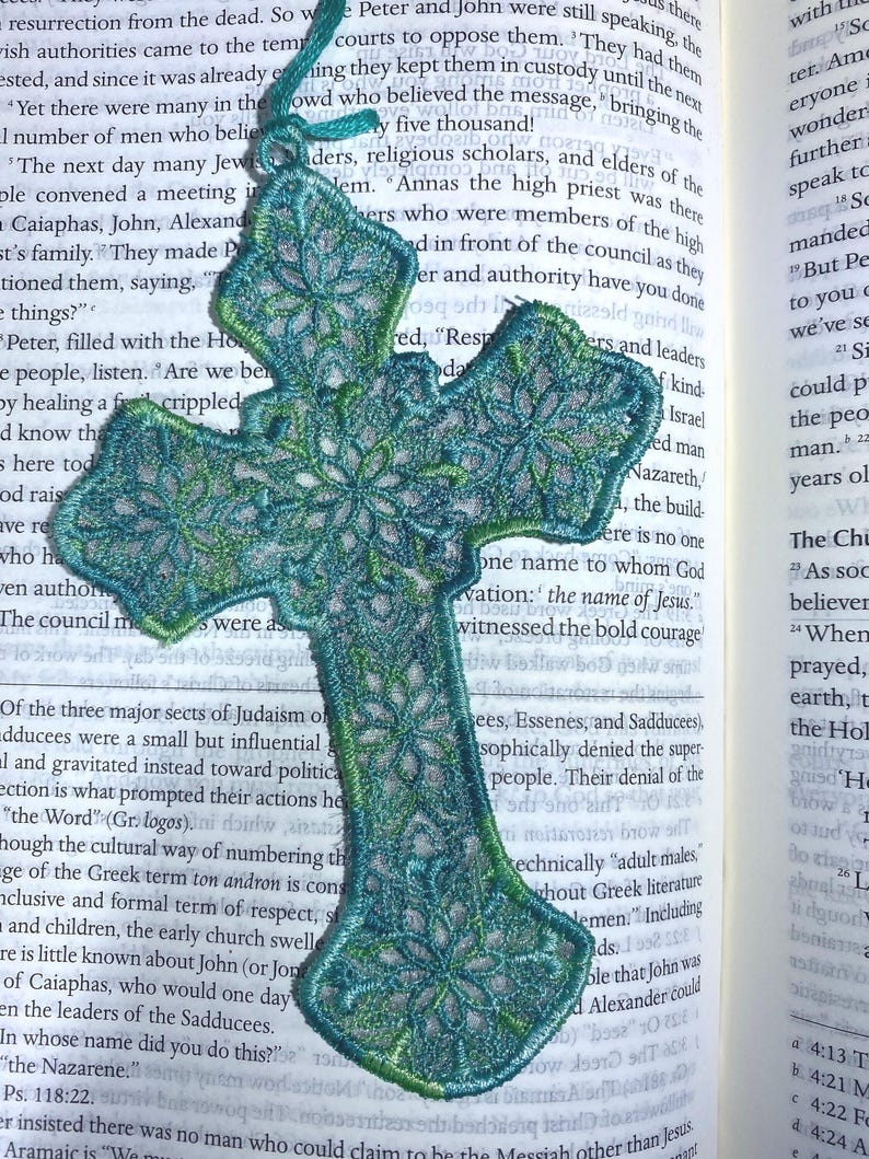 May include: Embroidered teal and green cross-shaped bookmark with floral detailing. The bookmark is made of a delicate, open-weave fabric and has a teal cord for easy placement in a book. The bookmark is resting on the pages of a book.