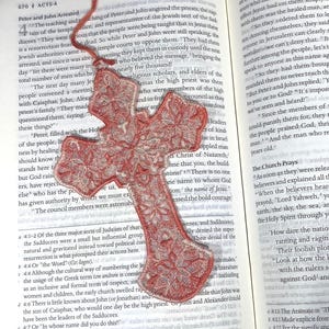 May include: Red cross-shaped bookmark with a floral lace design, resting on an open book. The bookmark is made of a translucent material and has a red ribbon attached. The book pages display printed text.