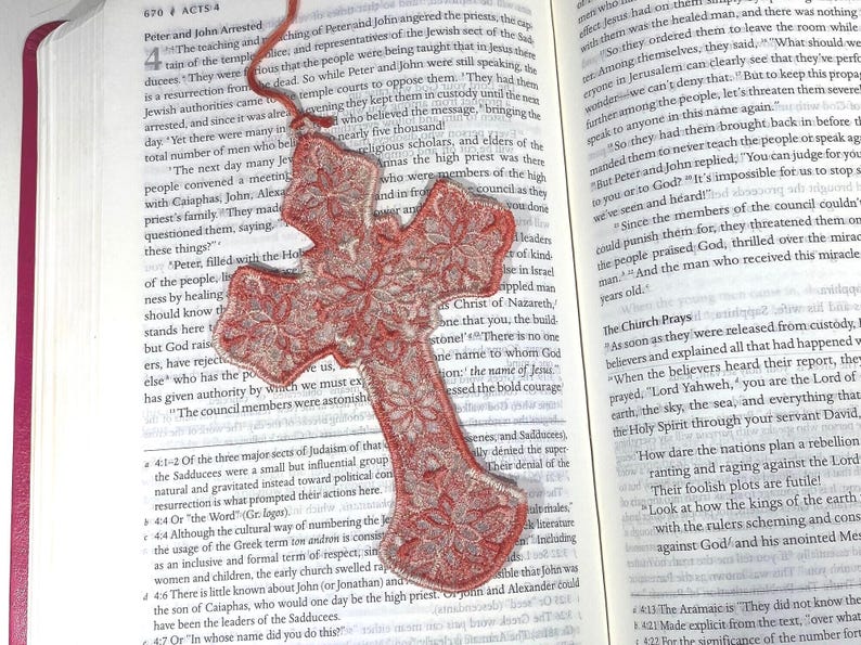 May include: Red cross-shaped bookmark with a floral lace design, resting on an open book. The bookmark is made of a translucent material and has a red ribbon attached. The book pages display printed text.