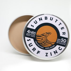 Puede incluir: Un contenedor de lata blanco con una etiqueta negra y naranja que dice "SUNBUTTER SURF ZINC SPF 50 ESPECTRO AMPLIO UV-A & UV-B 70g/2.5oz Pasta AUSTL 351659 4HRS RESISTENTE AL AGUA SEGURO PARA LOS ARRECIFES".