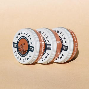 May include: Three white tins of Sunbutter Surf Zinc SPF50 sunscreen. The tins are stacked on top of each other. The tins are labelled with the brand name, product name, and SPF.