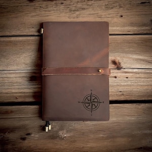 May include: A brown leather journal with a compass design engraved on the front cover. The journal has a leather strap closure with a brass button.
