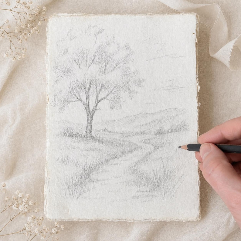 May include: A rectangular piece of textured, off-white paper featuring a graphite pencil drawing of a landscape. The sketch depicts a single tree in the foreground, a winding path leading toward rolling hills in the distance, and light clouds.