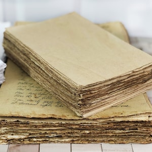 May include: A stack of rectangular, tan-colored paper sheets with rough, deckled edges. The top sheet is blank, while the sheet beneath it features handwritten cursive text in black ink. The paper has a textured, fibrous appearance throughout.