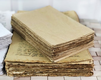 Pode incluir: Uma pilha de folhas de papel retangulares de cor castanha com bordas ásperas e irregulares. A folha superior está em branco, enquanto a folha abaixo apresenta texto manuscrito em cursivo com tinta preta.