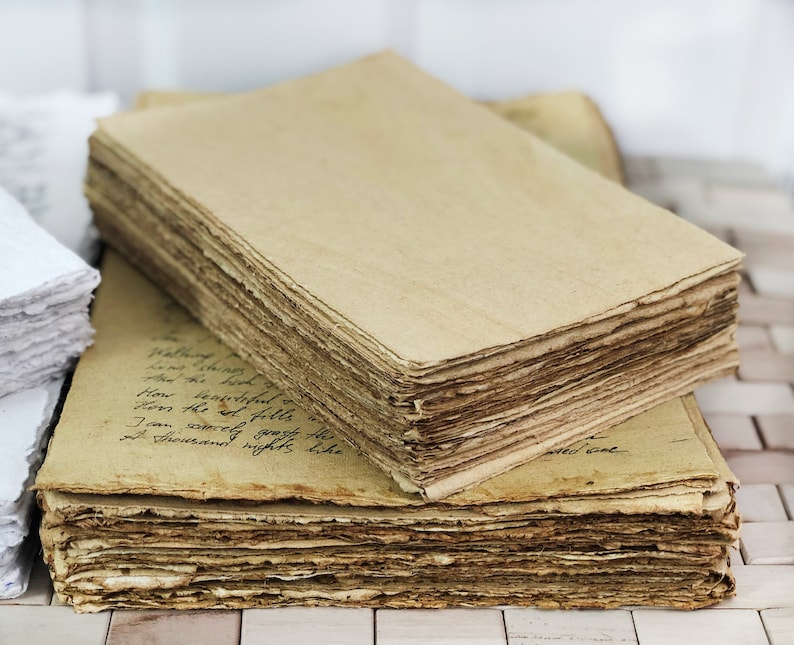 May include: A stack of rectangular, tan-colored paper sheets with rough, deckled edges. The top sheet is blank, while the sheet beneath it features handwritten cursive text in black ink. The paper has a textured, fibrous appearance throughout.