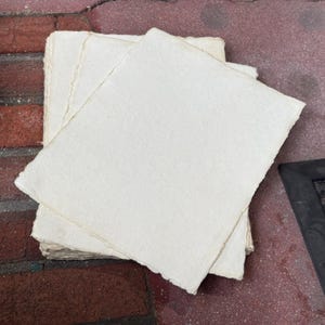 May include: A stack of rectangular sheets of off-white handmade paper with uneven, deckled edges. The paper has a visible fibrous texture and a matte finish, with each sheet layered slightly offset to reveal the rough, textured borders of the stack.