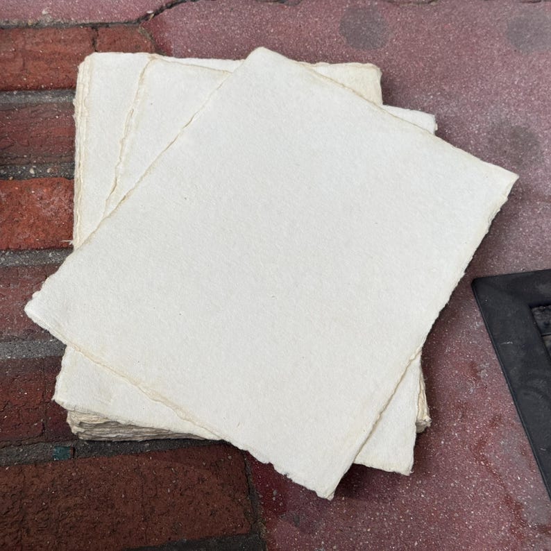 May include: A stack of rectangular sheets of off-white handmade paper with uneven, deckled edges. The paper has a visible fibrous texture and a matte finish, with each sheet layered slightly offset to reveal the rough, textured borders of the stack.