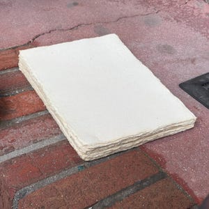 May include: A stack of rectangular, off-white paper sheets with a textured surface and uneven, deckled edges. The paper has a fibrous, handmade appearance with visible variations in the thickness and alignment of the stacked sheets.