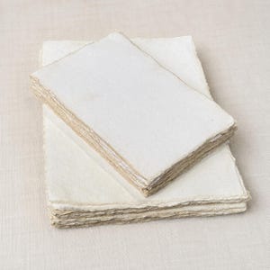 May include: A stack of rectangular sheets of off-white handmade paper with rough, deckled edges. The paper has a fibrous, textured surface and is layered in two distinct piles, showcasing the uneven, natural edges of each individual sheet.