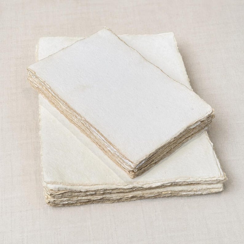 May include: A stack of rectangular sheets of off-white handmade paper with rough, deckled edges. The paper has a fibrous, textured surface and is layered in two distinct piles, showcasing the uneven, natural edges of each individual sheet.