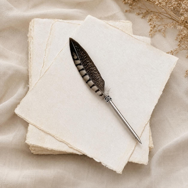 May include: A stack of square, off-white paper sheets with rough, deckled edges. Resting on top is a ballpoint pen featuring a brown and black striped feather quill attached to a silver-toned metal barrel with intricate engraved detailing.