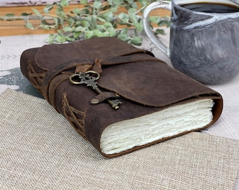 Op de afbeelding: Een bruin lederen gebonden dagboek met een sleutel aan de omslag. Het dagboek is gesloten en ligt op een beige stoffen oppervlak.