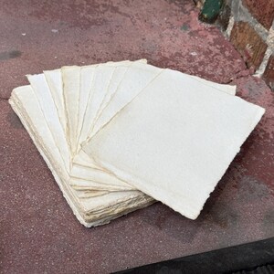 May include: A stack of rectangular off-white paper sheets with rough, deckled edges. The paper has a visible fibrous texture and is arranged in a fanned-out pile, showcasing the uneven, handmade quality of the edges on each individual sheet.