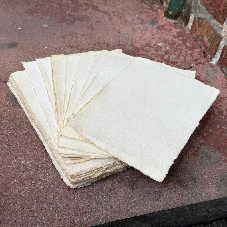 May include: A stack of rectangular off-white paper sheets with rough, deckled edges. The paper has a visible fibrous texture and is arranged in a fanned-out pile, showcasing the uneven, handmade quality of the edges on each individual sheet.