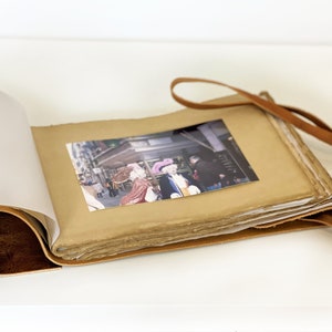 May include: A brown leather-bound journal with a strap closure. The journal is open to reveal a photograph of people in a street scene. The journal has a blank page on the left side.