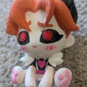以下が含まれることがあります： オレンジ色の髪、黒い猫の耳、赤い目をした3Dプリントの人形。人形は白い顔にピンクの頬と胸にピンクのハートがあります。人形の足は白く、ピンクの足跡が付いています。