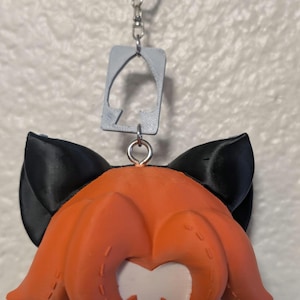 以下が含まれることがあります： オレンジ色の髪、黒い猫耳、赤い目をしたアニメ風のキャラクターが特徴の3Dプリントキーホルダー。キーホルダーには、灰色のスペード型のチャームと、取り付け用のシルバーのキーリングが含まれています。