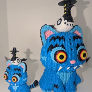 May include: Two blue cat figurines with black stripes and yellow eyes. Each cat has a small bird wearing a black hat perched on its head. The cats have white fangs and pink noses. One cat is larger than the other.
