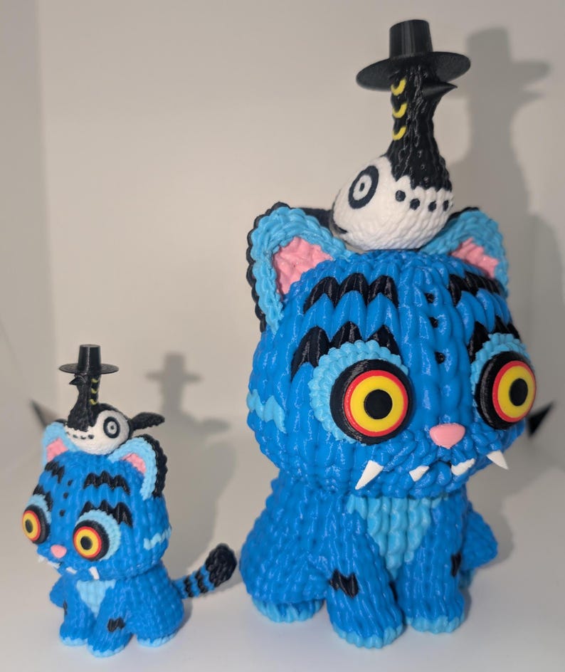 May include: Two blue cat figurines with black stripes and yellow eyes. Each cat has a small bird wearing a black hat perched on its head. The cats have white fangs and pink noses. One cat is larger than the other.