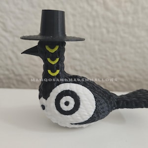 May include: A black and white bird figurine wearing a small black hat. The bird has yellow markings on its neck and a large white eye with a black center. The text "MANGOSANDMARSHMALLOWS" is visible.