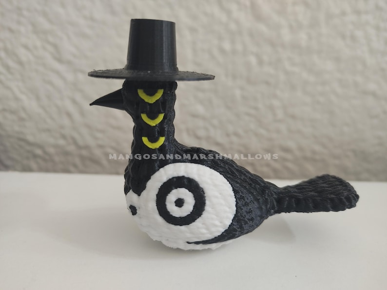 May include: A black and white bird figurine wearing a small black hat. The bird has yellow markings on its neck and a large white eye with a black center. The text "MANGOSANDMARSHMALLOWS" is visible.