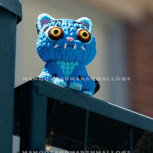 May include: A blue crocheted cat figurine with large yellow eyes, white fangs, and the text "MANGOSANDMARSHMALLOWS". The cat is perched on a dark blue metal railing. The cat's fur is a vibrant blue.