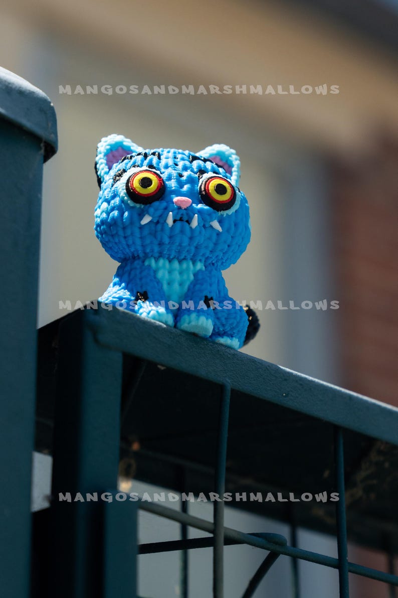 May include: A blue crocheted cat figurine with large yellow eyes, white fangs, and the text "MANGOSANDMARSHMALLOWS". The cat is perched on a dark blue metal railing. The cat's fur is a vibrant blue.